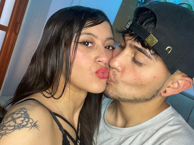 A selfie of ScarlettAndAlan and another person kissing. ScarlettAndAlan, a woman with long black hair, a nose ring, and a floral tattoo on her left shoulder, has full lips and is wearing a black lace top. The other person, a man with dark hair under a black cap, is kissing her. He's wearing a gray shirt. Both have closed eyes and are facing the camera. The man has a slight beard and a small nose. The woman's lips are pink and slightly parted. The man's cap has a small gold logo. The woman's makeup is subtle.