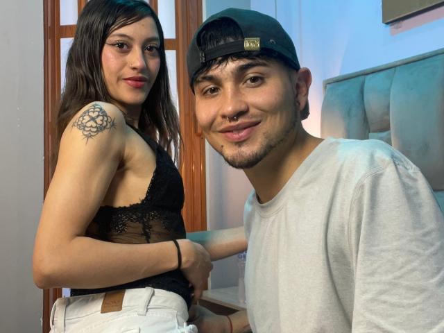 ScarlettAndAlan and Alan are in a photo. Scarlett has long black hair, red lipstick, and a black lace tank top with white pants. She has a floral tattoo on her right shoulder. Alan has a black cap, nose ring, and a gray shirt. Both are smiling. Scarlett's right hand rests on Alan's shoulder. The image is clear with natural light highlighting their faces. Alan's left ear is pierced. The photo is casual and intimate.