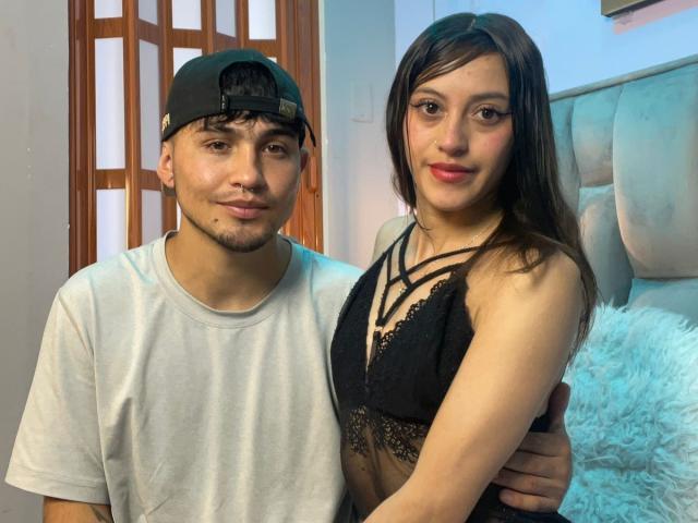 ScarlettAndAlan, with brown hair and black lace bra, stands arm around Alan, in a white tee and black cap, both smiling, in a bedroom. Alan has a beard.