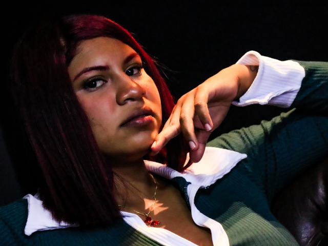 DominFerrari, a young woman with medium brown skin and straight, dark auburn hair, leans on a leather chair. She wears a green rib-knit sweater with a white collar and a necklace with a red pendant. Her right hand rests on her chin, and she gazes slightly to the right with a neutral expression. The lighting is dark, highlighting her face and hair.