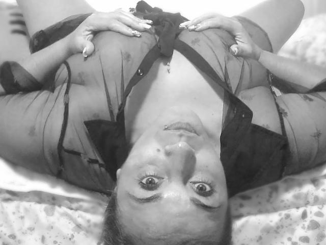 Black and white photo of ZoeHotXX lying on a bed, head upside down. She has a light skin tone, small nose piercing, and visible freckles. Her wide-eyed, slightly open-mouthed expression looks surprised. She wears a sheer black robe with dark straps, hands gripping the robe near her chest. Her nails are painted with a floral design. The bed has a patterned sheet. The photo is tightly framed, focusing on her face and upper body. The texture of the robe is slightly transparent, showing her skin underneath. Her eyebrows are slightly raised.