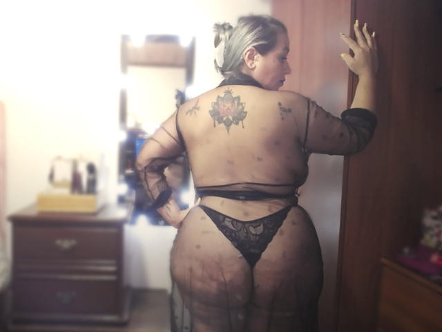 ZoeHotXX stands with her back to the viewer, right hand on a wooden door. She's wearing a sheer black robe, exposing her tattooed back and black lace thong. Her blonde hair is tied up. A floral tattoo is centered on her upper back. She has a curvy, slightly overweight build. The room has a wooden dresser with various items on it. The lighting is soft, creating a slightly blurred effect. The image has a sensual, provocative tone.