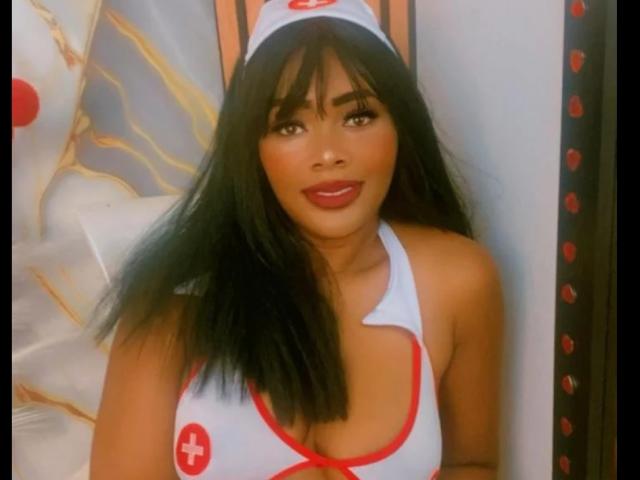 AstridDaviz, a Latina woman with long black hair and bangs, wears a white nurse costume with red trim and a red cross. She has red lipstick and a confident smile. Her costume has a deep neckline, revealing cleavage. She wears a matching nurse cap. The image is slightly blurred. The costume's red cross is on the left side of her chest. The lighting is warm.