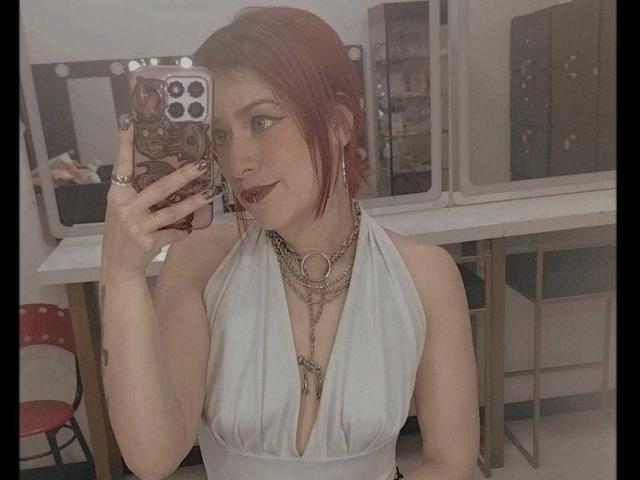 LolyYunno with red hair, white halter dress, deep cleavage, chains, red lipstick, holding phone, mirror selfie, visible tits, cute, small tattoo on left arm.