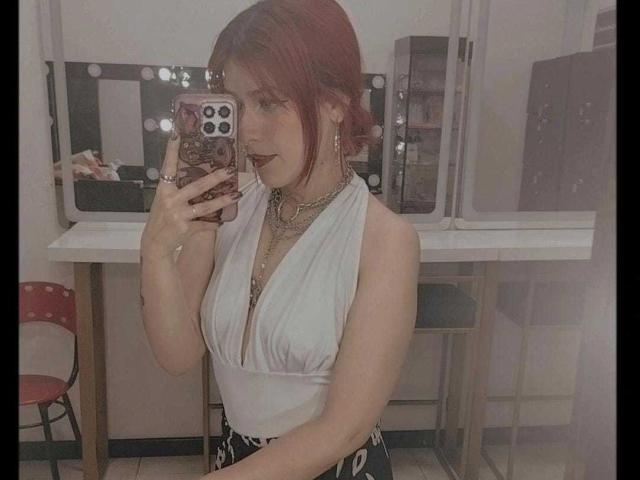 LolyYunno with red hair, white halter top showing tits, black pants, holding phone, mirror selfie, dark lipstick, gold jewelry, chair in background, white wall.