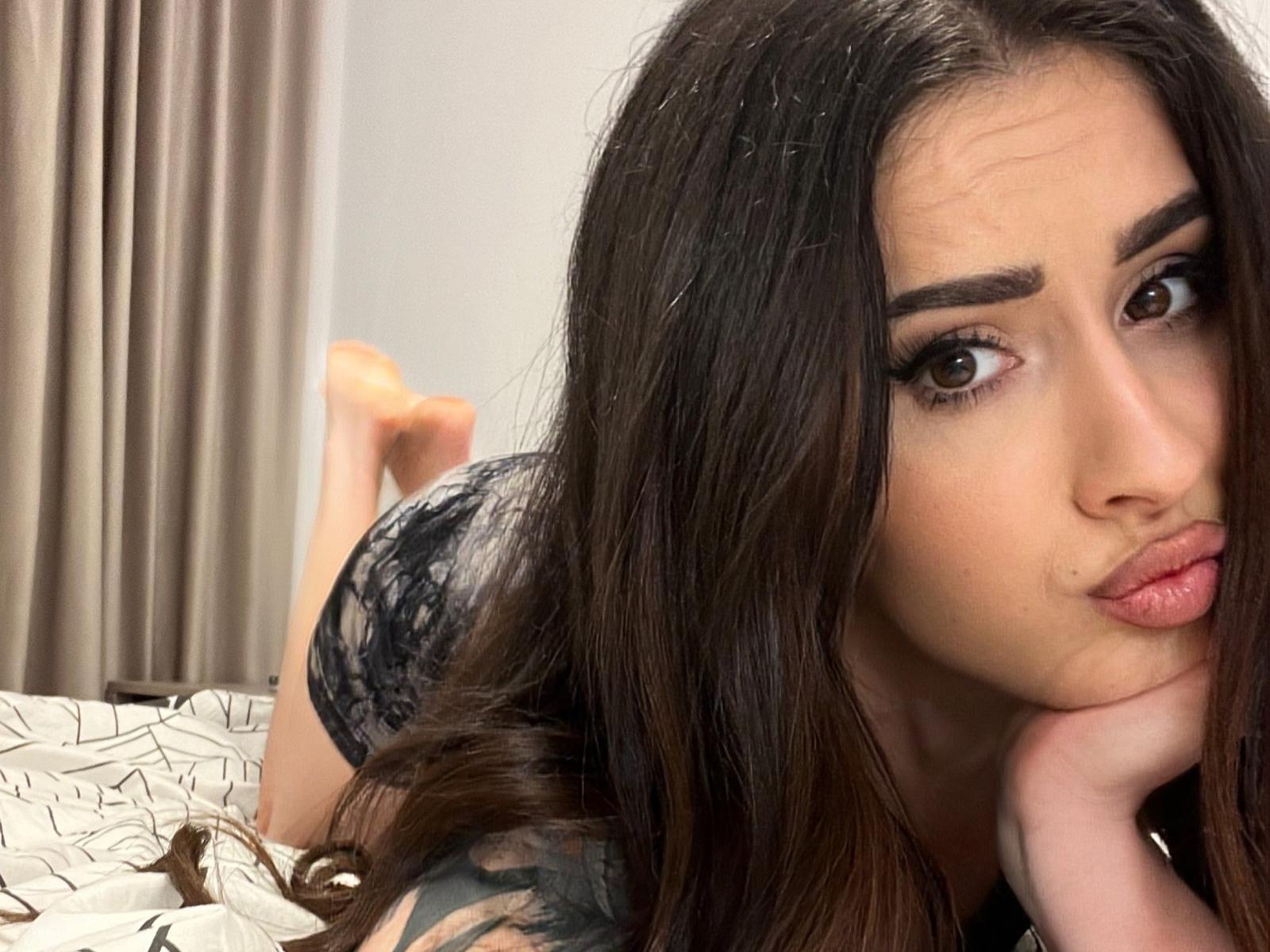 AriaAdda with long brown hair, black lace top, pouty lips, and dark eyeliner, lying on bed, hand under chin, looking at camera, bare feet in background.
