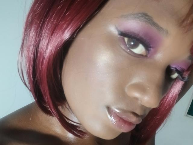 Close-up of CristalGonzales with dark brown skin and shiny, smooth complexion. She has bright red, shoulder-length hair that's slightly parted to the left. Her eyes are large with prominent black eyeliner and pink eyeshadow extending to her upper eyelids. Her lips are glossy and slightly parted. The light highlights her cheekbones and upper lip, creating a shiny effect. CristalGonzales is looking to the right with a subtle, relaxed expression. The image is sharp with a slight shadow on the right side of her face.