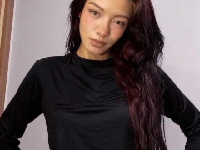 IvorySelene with long, dark red hair, wearing a black long-sleeve shirt, hands on hips, looking slightly to the side with a neutral expression.
