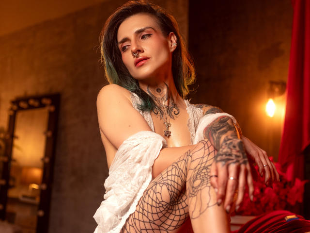 EmaLeBlanc, a pale-skinned woman with green-tinted black hair, sits with a confident, slightly defiant expression. She has multiple tattoos on her neck, chest, and hands, including a spiderweb on her right hand and a large, intricate design on her left arm. She wears a white lace shirt, partially off her shoulders, and fishnet stockings. Her left leg is bent, exposing more tattoos. Her nose is pierced with a small ring. Warm, orange lighting from the background creates a dramatic, almost sensual atmosphere. Red curtains are visible in the background. The image has a modern, edgy, and slightly rebellious style.
