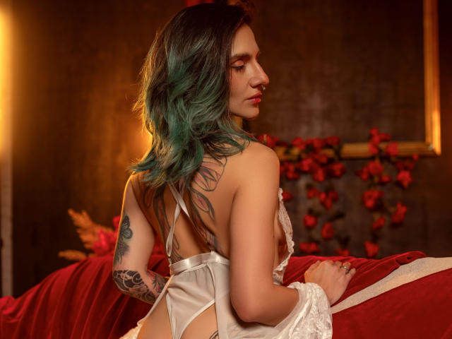 EmaLeBlanc with teal-green hair, white lace-up lingerie, back tattoo, and arm tattoo, sitting on red bed, looking over shoulder, showing her tits and ass. Warm, lit-up room.