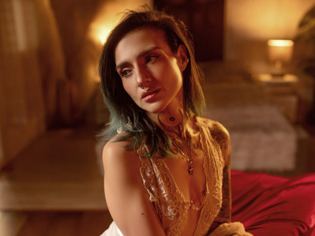 EmaLeBlanc has medium-length, dark greenish-blue hair and is looking slightly to the left with a subtle, pensive expression. She has a nose ring in her right nostril and a tattoo on her left side of the neck. She's wearing a gold, lace, low-cut top that reveals part of her cleavage. The lighting is warm, casting a soft glow on her light skin. The background is blurred, focusing attention on her. She has a tattoo on her right upper arm. The image has a slightly warm, brownish tone. Her makeup is subtle, emphasizing her eyes. The red bed in the background adds a contrasting color.