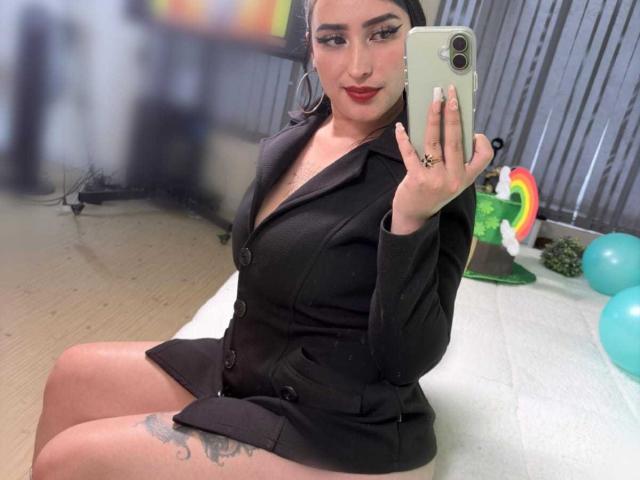 Chloey69 mit rotem Lippenstift, einem schwarzen Blazer und Shorts, macht ein Selfie mit ihrem Handy und zeigt dabei ihren Hintern und ein Tattoo an ihrem Oberschenkel, während sie auf einer weißen Oberfläche sitzt..