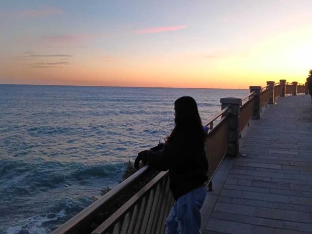 EmmaMae stands on a wooden boardwalk with a metal railing, facing a calm ocean during sunset. She wears a black hoodie and blue jeans, with long hair covering her face. The sky is a gradient from orange to pink, reflecting on the water. The boardwalk extends to the right, with wooden posts and small lights. The ocean has gentle waves, and the horizon is visible. The composition uses natural light and emphasizes the serene, coastal scene.