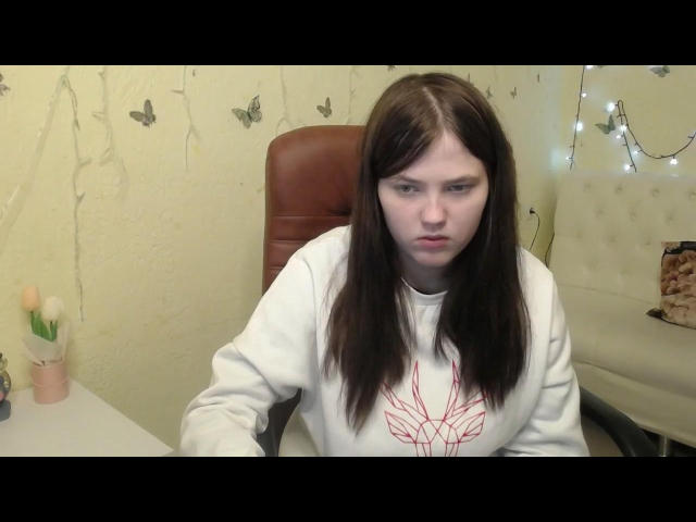 KateSii, with long brown hair and white sweater, sits in a brown leather chair, looking annoyed. She's got small tits and a serious face. A table with flowers is nearby.