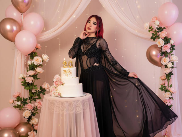 MiilaRouge with red hair, black sheer dress, and long sleeves, standing by a white cake with "20" on it, surrounded by pink and gold balloons, roses, and fairy lights.