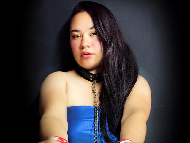 LillyRous with long black hair, blue strapless top, black leather collar with chain, and red wristbands, looking slightly to the side with a neutral expression.
