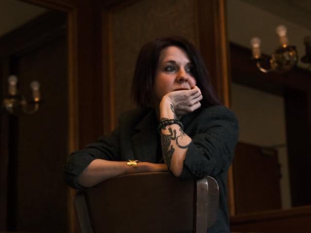 SherlineStone with dark hair, black jacket, and tattooed arm rests on brown chair, hand near mouth, looking thoughtful. Gold bracelet on wrist. Dim, warm lighting.