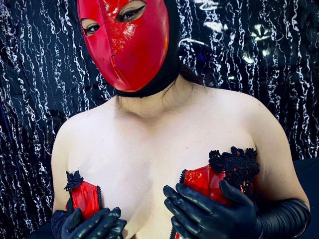 LillyRous with red latex mask, black lace gloves, holding red zipped-up tits with black lace trim, fair skin, dark eyes, against black and white textured background.