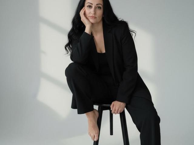 VioletNice with long black hair, sitting on a stool, wearing a black suit, barefoot, head resting on hand, smiling, black pants, white background, soft light.