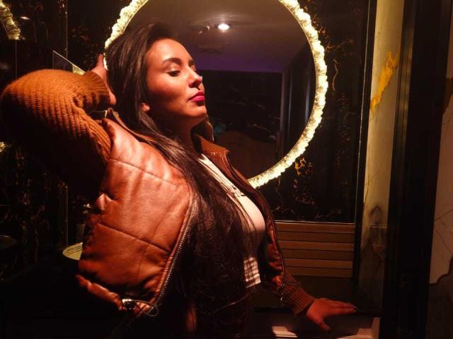 MelizaQueen stands in a dimly lit room, facing a round mirror with a glowing, beaded frame. She has long, straight black hair, and wears a brown leather jacket over a white top. Her right arm is raised, hand in her hair, and she has a relaxed, confident expression. Warm light highlights her face and jacket, creating a contrast with the darker background. The mirror reflects her profile and part of the room. The image has a candid, natural style.