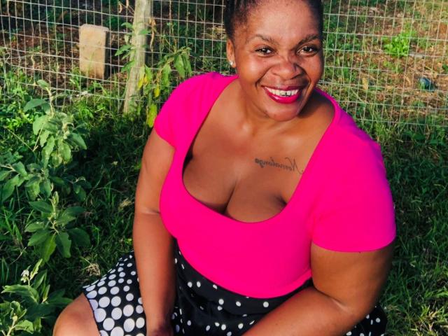 QueenBebe is a dark-skinned woman with a bright pink, low-cut top that shows her large breasts. She's sitting on grass with a black polka-dotted skirt, smiling at the camera. She has red lipstick and a tattoo just above her cleavage. Her hair is in a bun. The image is colorful and focused on her. She's surrounded by green plants and a wire fence.