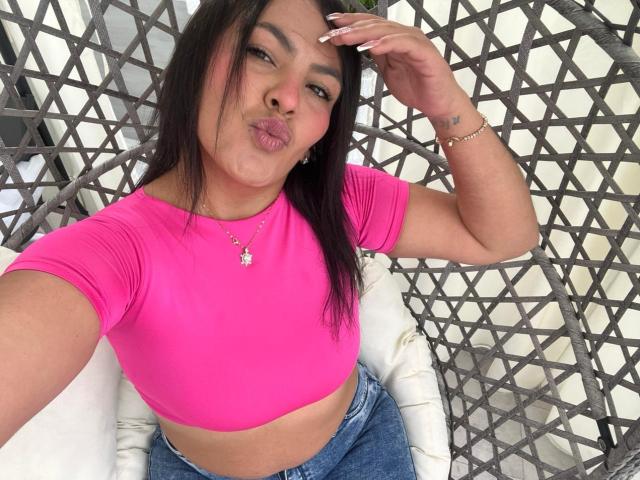 ScarlettOxford with medium tits, pink crop top, and blue jeans, pouting her lips, long dark hair, wearing a necklace and bracelet, sitting in a grey woven chair.