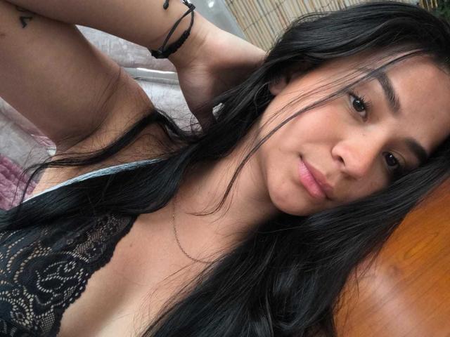 RouxiMori with long black hair, lace top, and a slight smile, showing her tits, arm tattoo, and black bracelet, lying on a bed. Her brown eyes are soft.