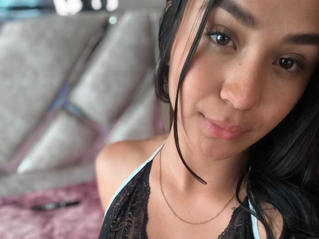 RouxiMori has a cute face with brown eyes, black hair, and a subtle smile. She's wearing a black lace top with a thin necklace, showing her tits.