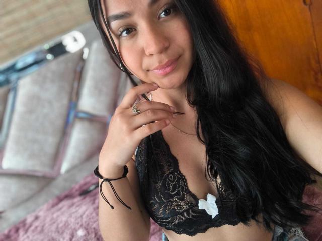 RouxiMori with long black hair, wearing a black lace bra with a white bow, smiling, touching her chin, ring on finger, bracelet, light brown eyes, pink lips.