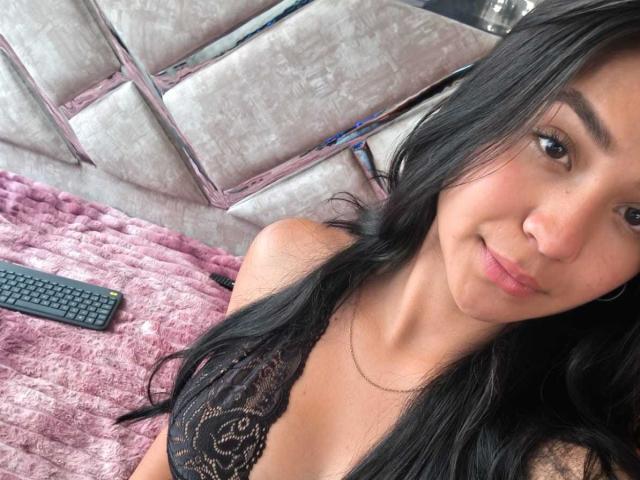 RouxiMori with long black hair, wearing a black lace bra, lying on a pink fuzzy blanket, remote control to the left, smiling at the camera, cute, sexy.