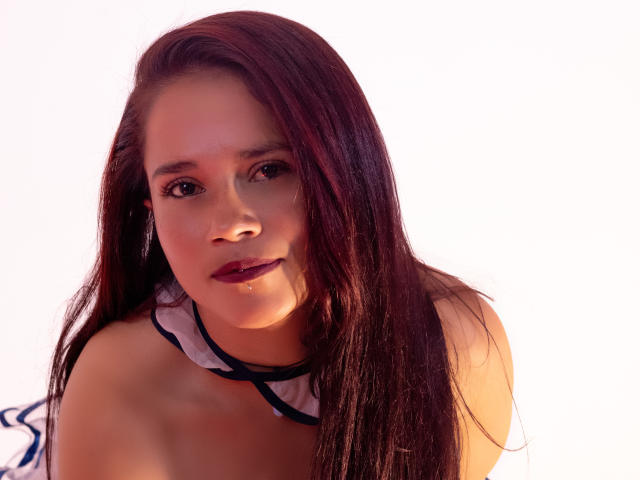 NicoleJewel has long brown hair, dark eyes, and pink lipstick. She's wearing a white top with blue trim, looking slightly to the side with a gentle smile.