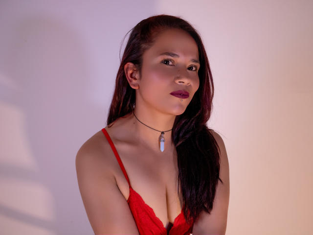 NicoleJewel has long, dark brown hair parted to one side. She wears a red lace bralette with thin straps, revealing her cleavage. A black choker with a small, blue teardrop pendant hangs around her neck. Her makeup is subtle but includes dark red lipstick. The lighting casts a soft shadow on her right side. Her skin is smooth and she looks slightly to the left with a neutral expression. The image has a warm, slightly purple-tinted light.