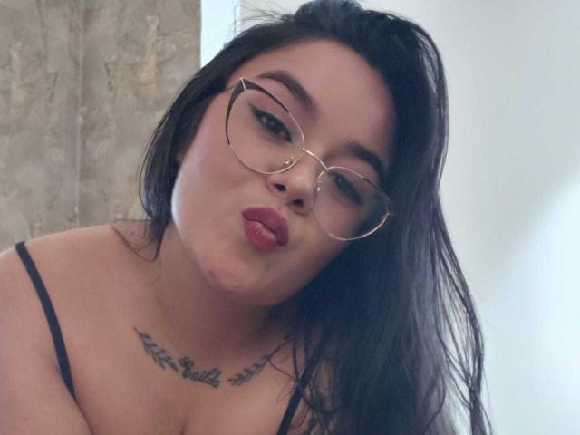 HannahMolly with pouty red lips, black straight hair, and glasses, showing a chest tattoo, wearing a black strap top, in a slightly tilted selfie.