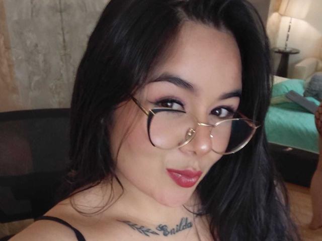 HannahMolly with black hair, glasses, and red lipstick, showing a tattoo under her chin, smiling at the camera. Her tits are slightly visible. Bedroom setting.