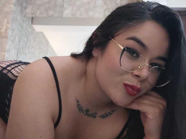 HannahMolly with glasses, red lipstick, black fishnet lingerie, and a tattoo under her tits, pouting and leaning on her hand. Her ass is visible in the background.