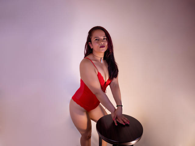 NicoleJewel with red lingerie, leaning on a stool, hands on top, wearing a necklace, dark hair, light skin, small tits, visible ass, slight smile, standing in a studio.