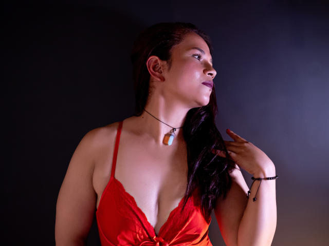 NicoleJewel with dark hair, red lace dress, small tits, light skin, pendant necklace, black bracelet, looking to the right, dark background, hand near face.