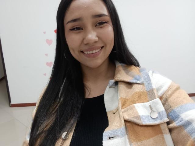 Karollteyes has long black hair, smiling directly at the camera. She wears a plaid jacket with beige, white, and blue stripes over a black ribbed top. Her skin is smooth and she has subtle makeup with defined eyebrows. She's in a brightly lit room with a whiteboard in the background. The whiteboard has small pink hearts. Her nose is slightly upturned, and she has a natural, confident look. The image is a selfie with a casual, approachable vibe.