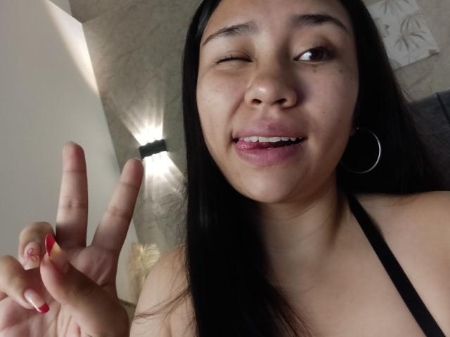Close-up photo of Karollteyes, a young woman with long black hair, winking with her right eye and smiling with slightly parted pink lips. She has medium brown skin, small dark eyebrows, and is wearing a black halter top. She holds up her right hand with fingers in a "peace" sign, showing red-painted nails. A silver hoop earring is visible in her left ear. The background is blurred, with a light-colored wall and a lamp.
