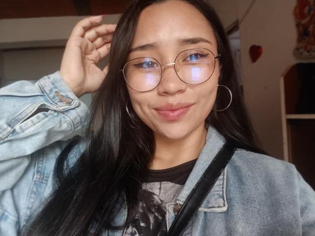 Karollteyes has long black hair and wears large, round glasses with a thin frame. She has light brown skin and blue eyes. She's dressed in a light blue denim jacket over a black graphic t-shirt. Her right hand is raised, touching her hair. She wears small silver hoop earrings. A black strap crosses her chest. The background is blurred, featuring a red heart on a white wall. The image has a natural, slightly overexposed light. Karollteyes has a subtle, confident smile.