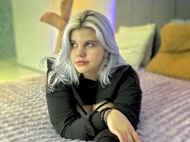 JoannLowe with pale blue hair, black top, and black bracelet, lying on bed, legs up, looking off to side, soft pink bed, yellow pillow, subtle purple light.