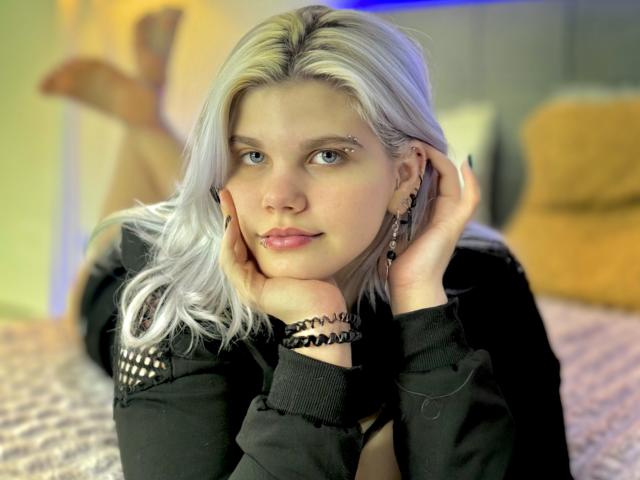 JoannLowe with platinum blonde hair, black shirt, and earrings, lying on a bed, hands on cheeks, legs up, blue light in background, looking at camera, slight smile.