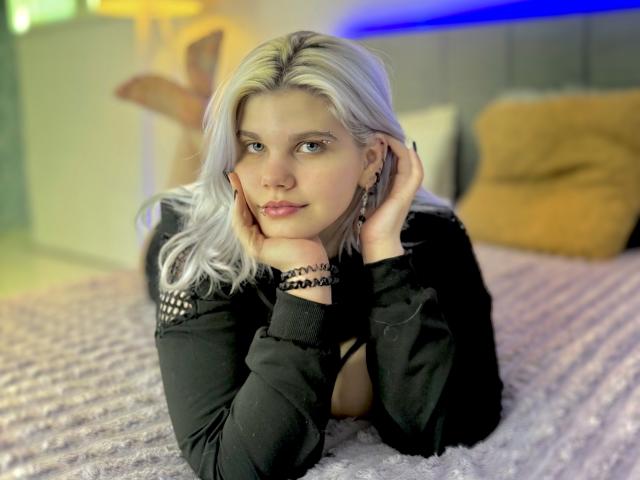 JoannLowe with platinum blonde hair, black top, laying on a bed, hands on cheeks, blue LED strip in background, pink textured blanket, yellow pillow, and black bracelets.