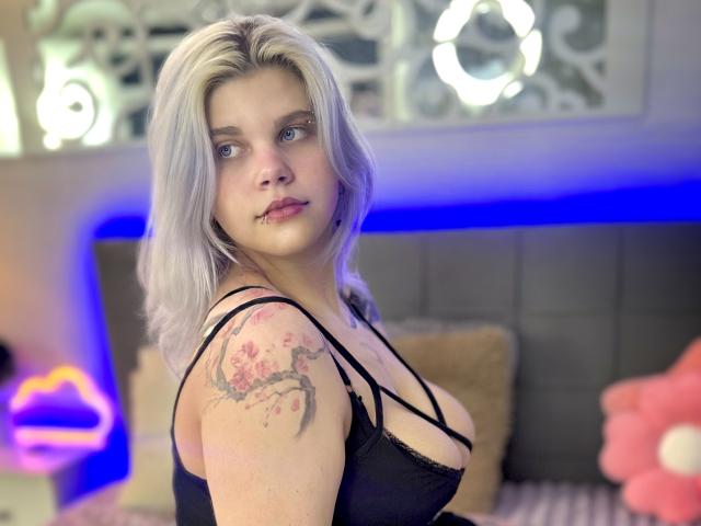 JoannLowe with platinum blonde hair, black lace top, showing her big tits and tattooed arm, looking over her shoulder, blue neon lights in background.