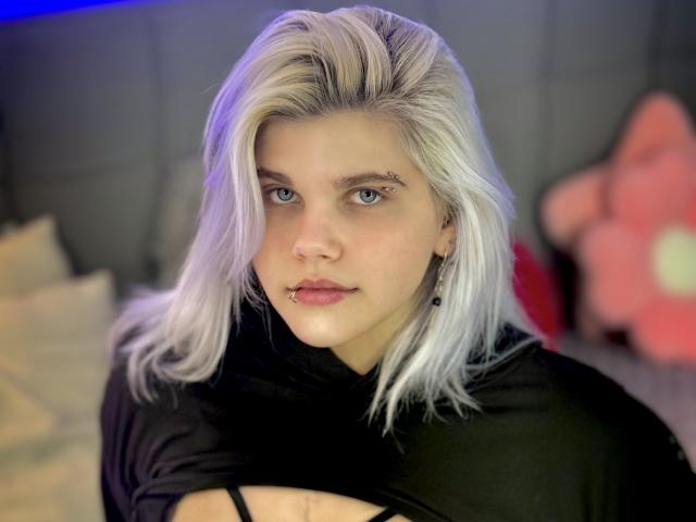 JoannLowe has pale skin, platinum blonde hair, blue eyes, a nose piercing, and a lip piercing, wearing a black top, with a serious expression, in a softly lit room.