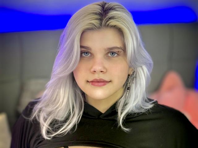 JoannLowe with pale skin, platinum blonde hair, blue eyes, black hoodie, nose piercing, slight smile, blue neon light in background. Boobless, no visible ass, no visible pussy.