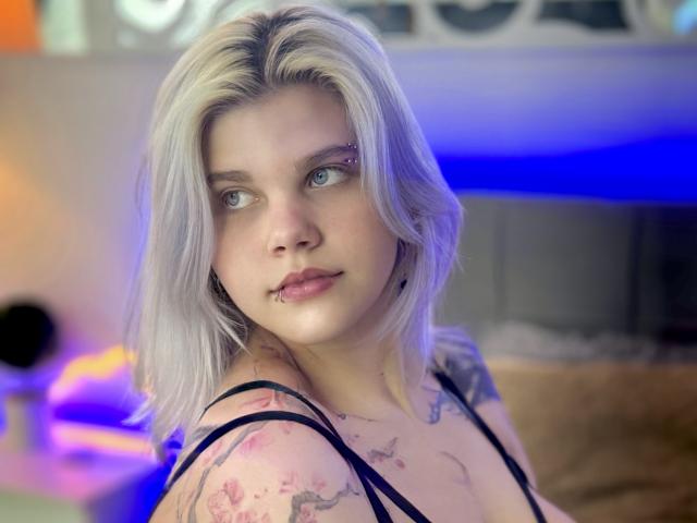 JoannLowe with blonde hair, blue eyes, and tattoos, wearing a black strappy top, has a nose piercing and a slight lip piercing, looking to the side with a subtle smile.