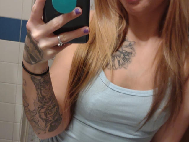 Lexxx444, avec de longs cheveux blonds, portant un débardeur bleu clair, montrant des tatouages sur sa poitrine et son bras, tenant un téléphone avec des ongles violets, dans un autoportrait devant un miroir de salle de bain..