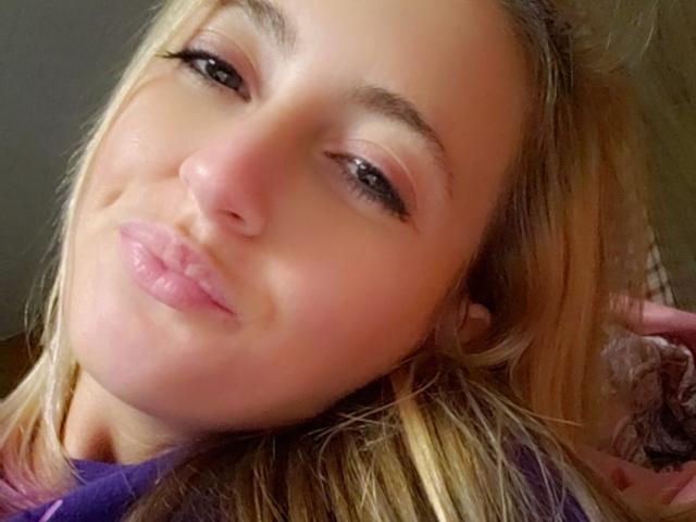 Lexxx444 a un visage mignon avec des cheveux blonds, des lèvres roses et un maquillage discret. Son nez est légèrement retroussé. Elle regarde l'appareil photo avec un petit sourire joueur..