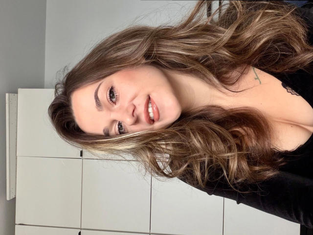 Minnie-hot has long, wavy brown hair, light skin, and a cute smile. She's wearing a black top, showing some cleavage, with a small tattoo on her left chest.